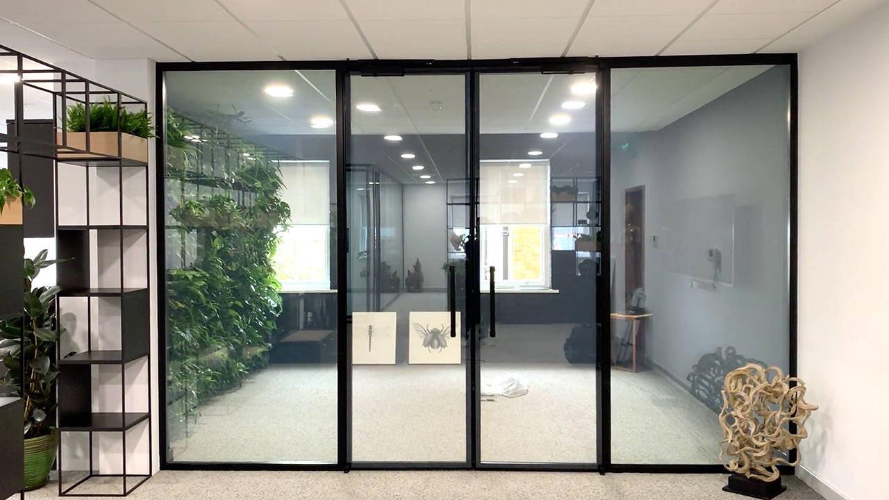Black-framed glass office partition with double doors in the clear state, a green living wall visible behind