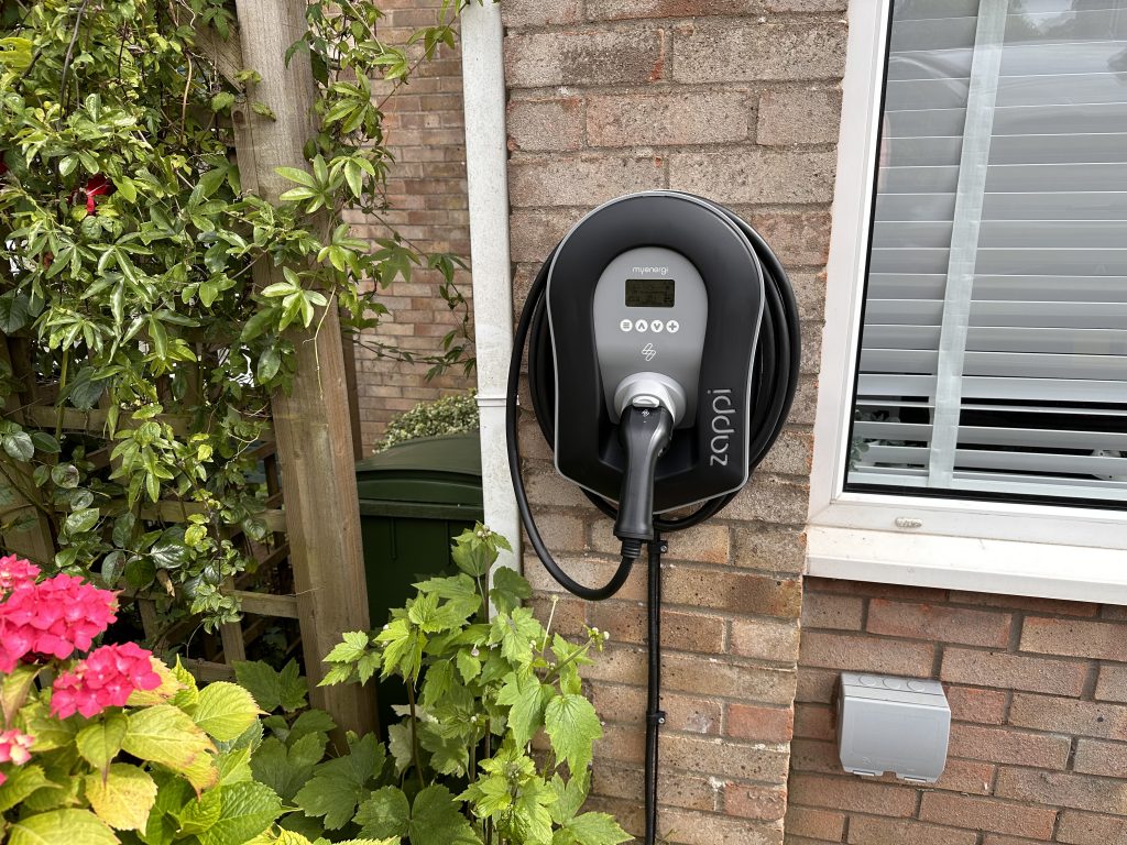 A wall-mounted home EV charger installed on a brick house among garden plants